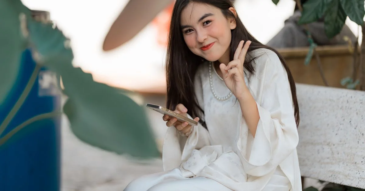 Jeune femme souriante en robe blanche, posant avec un smartphone à l'extérieur et faisant le signe de la paix.